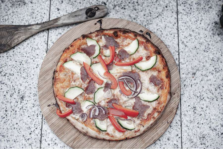 Rezept Foto Holzofen Pizza Komplettanleitung. Fertig gebackene Pizza mit Salami und frischem Gartengemüse auf einem Holzbrett angerichtet.