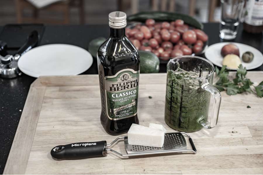 Rezept Foto Kräuter und Garten Pesto. Schneidebrett mit Olivenöl Flasche einer Parmesan Reibe und den Kräutern darauf.