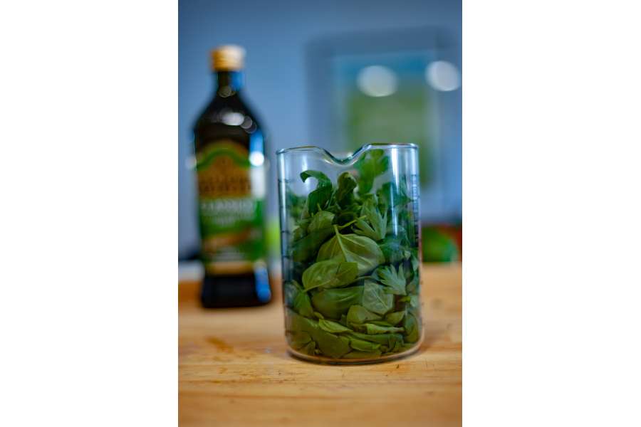 Rezept Foto Kräuter und Garten Pesto. Messbecher mit den Zutaten und dahinter steht eine Flasche natives Olivenöl.